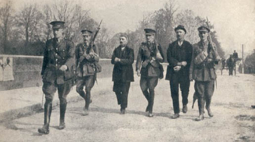 Black-and-white photograph of two men walking under military escort on a road
